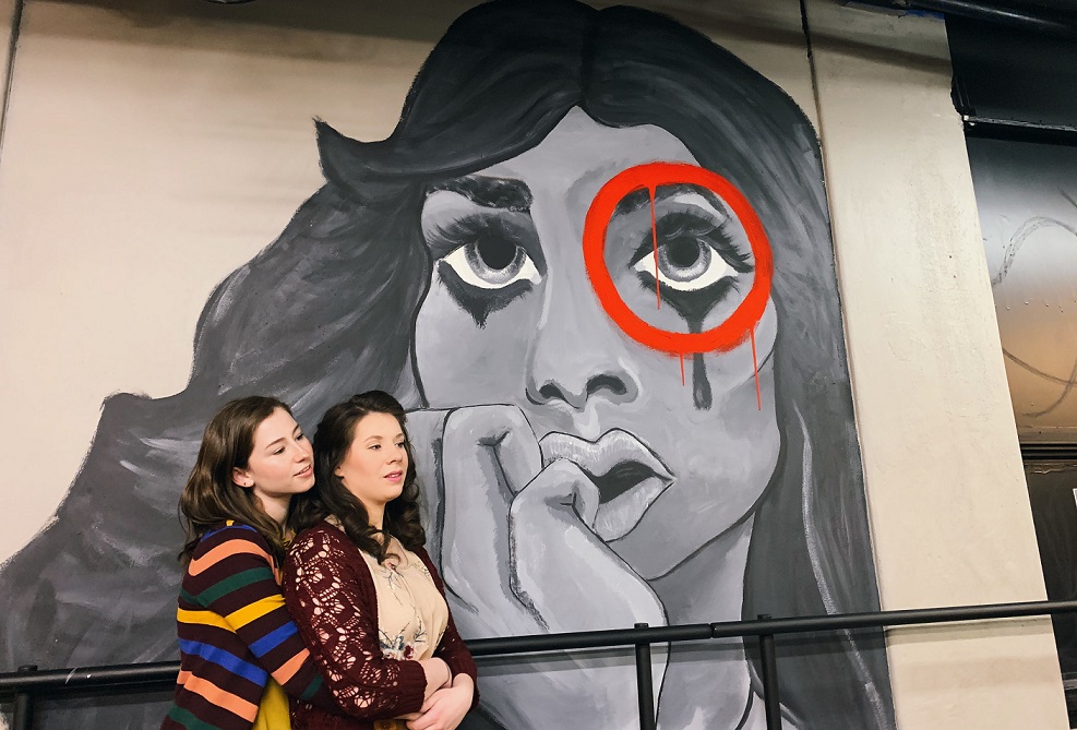 Two actresses pose in front of a picture of a crying woman for the Department of Theatre's production of Stop Kiss.
