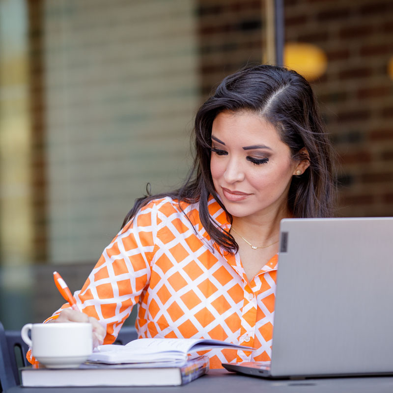 Student studying outdoors at a table with a laptop, open notebook, pen, and coffee cup while wearing an orange and white checkered top.