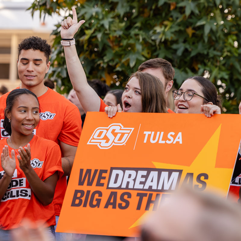 Excited OSU students holding "We Dream As Big As The Sky" sign with OSU-Tulsa branding at campus event.