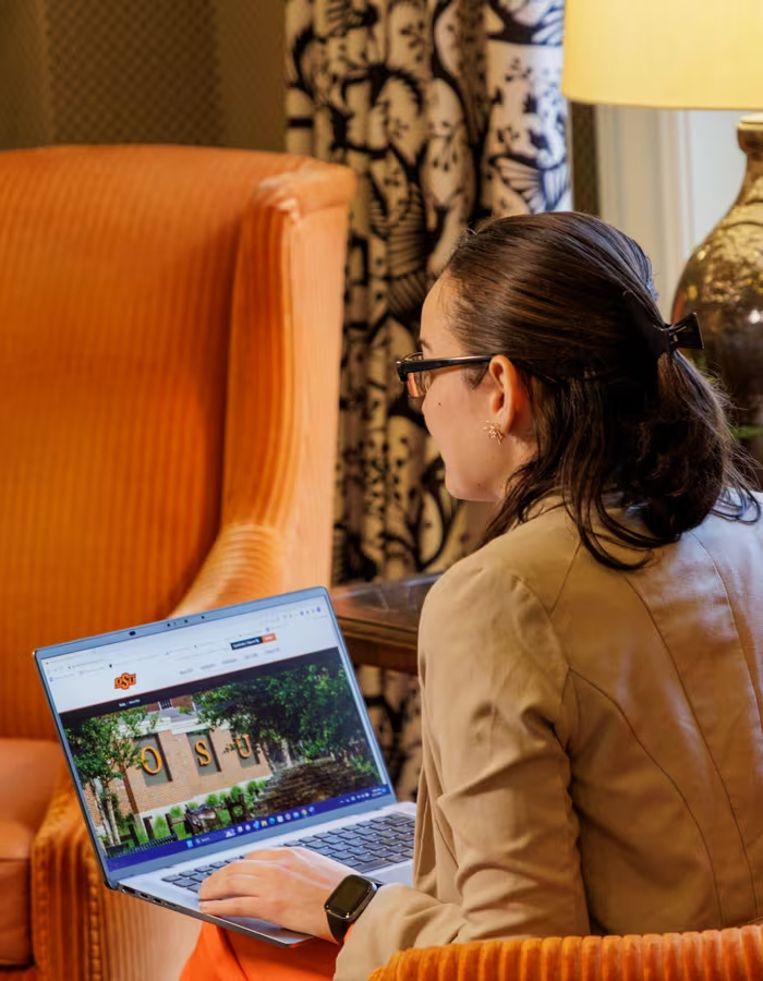 Student on laptop while sitting in oversized orange chair. Over the student's shoulder we see the browser is open to an About OSU webpage.