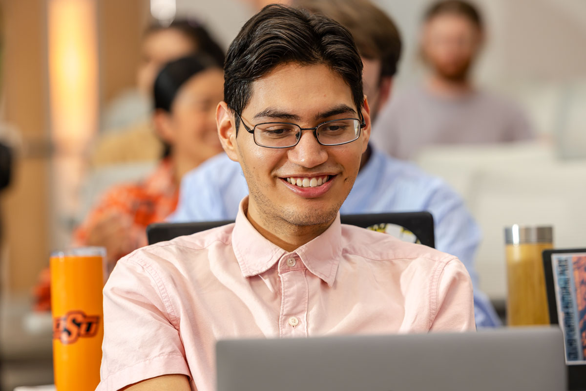 A male student wearing glasses smiles while working on a laptop in a computer lab.