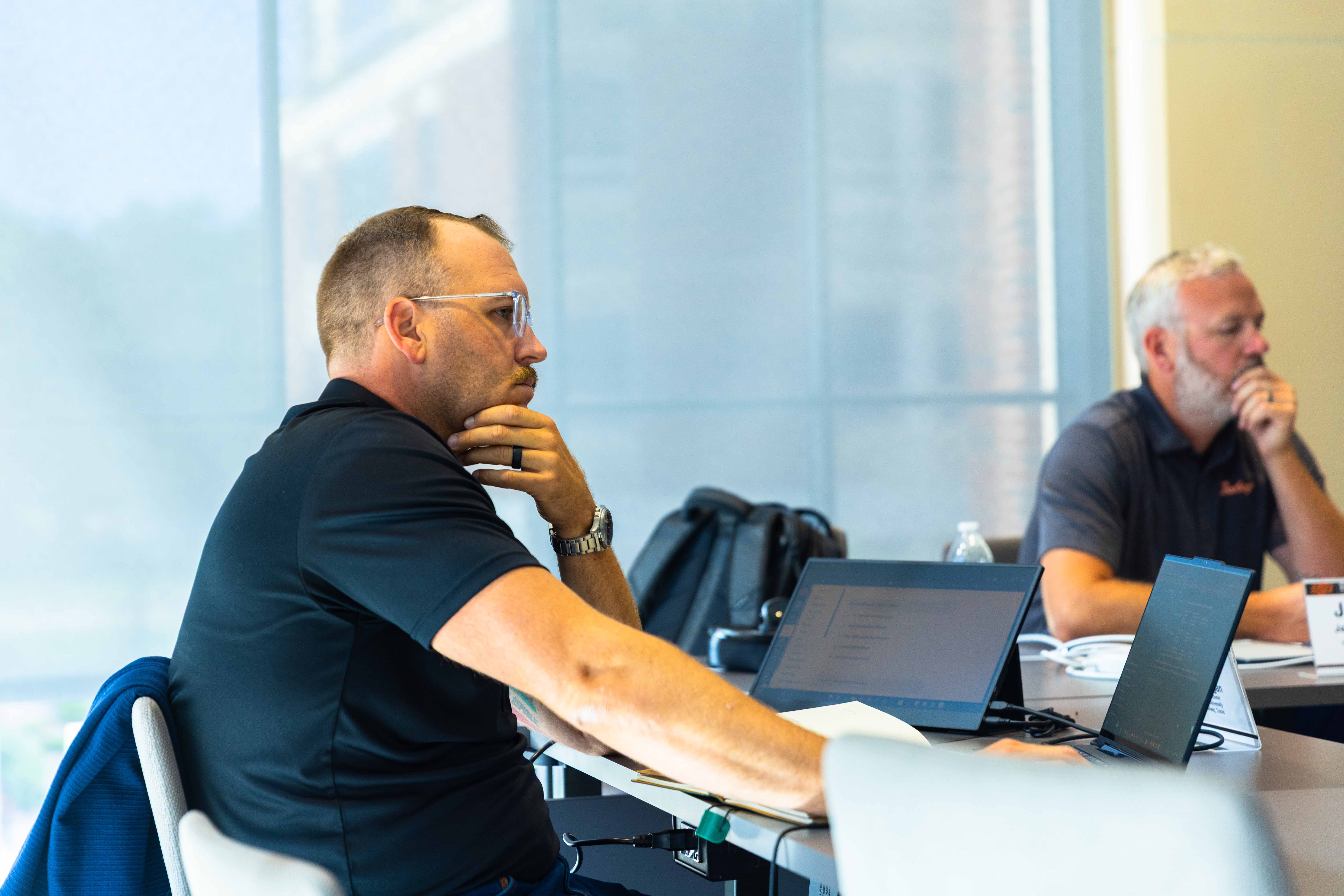 A man sits at a conference room table with a laptop looking intently at a speaker.
