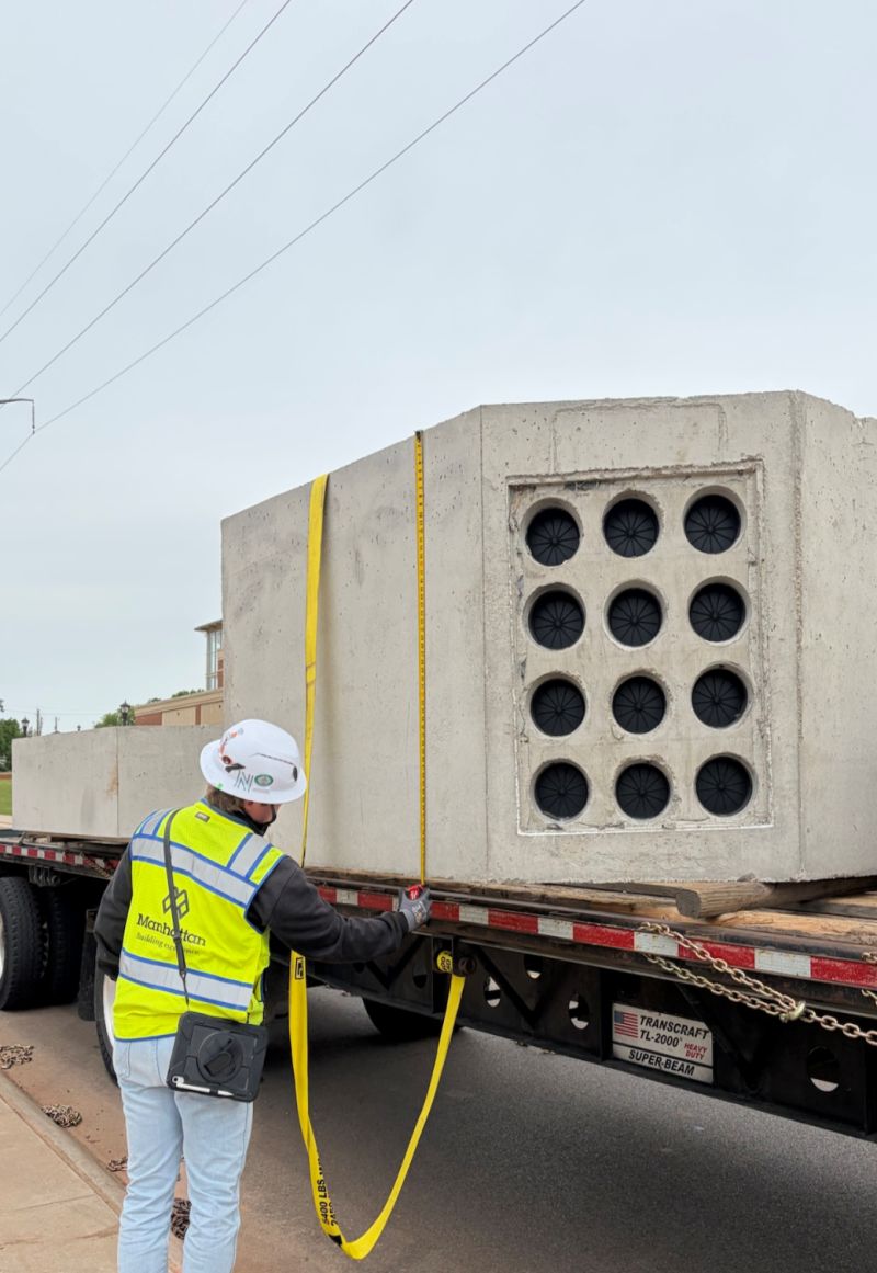 Construction worker in a high-vis vest measures a large precast concrete utility structure secured on a flatbed trailer.