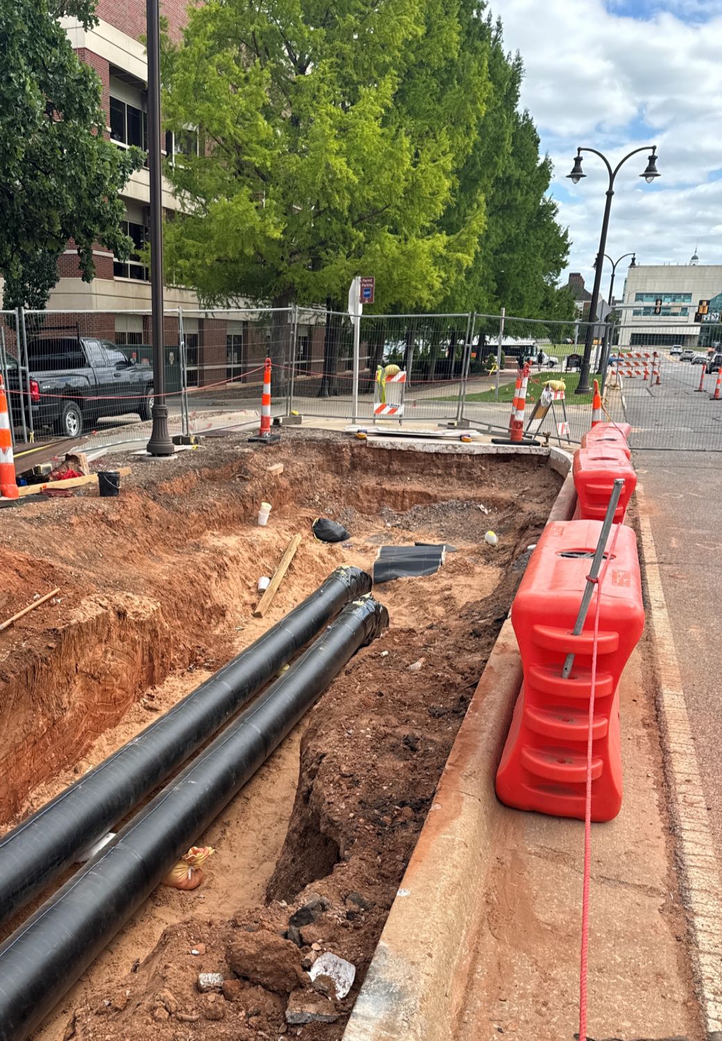 Open trench along a city street with large black utility pipes installed underground, surrounded by safety fencing and traffic barriers.