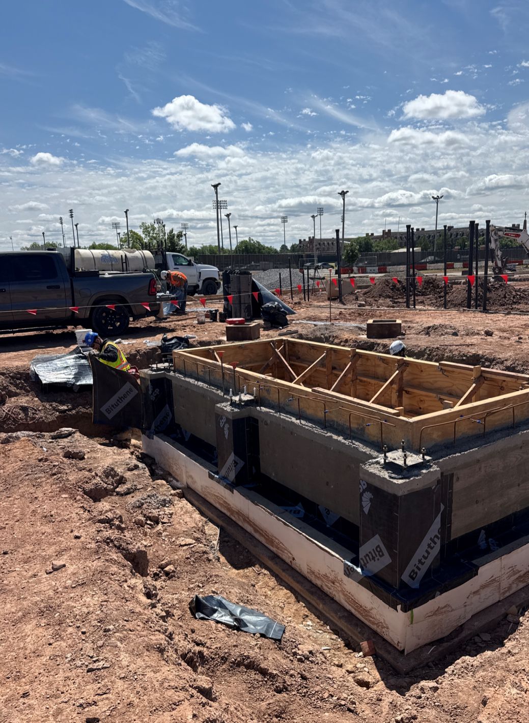 Concrete elevator pit foundation under construction at a work site, with formwork, waterproofing, and excavation visible around the structure.