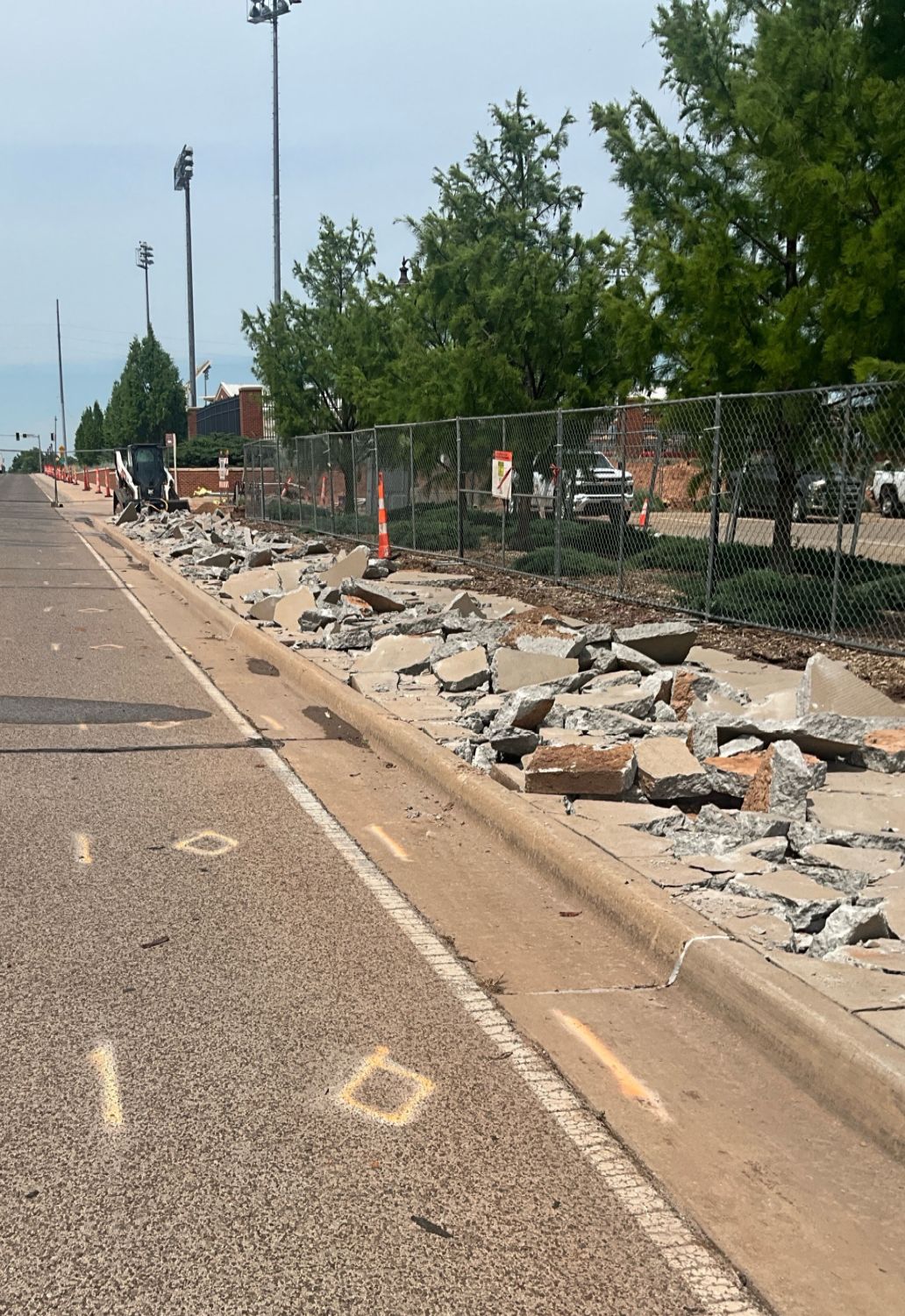 Roadside construction showing demolished sidewalk and curb sections alongside a temporary fence.