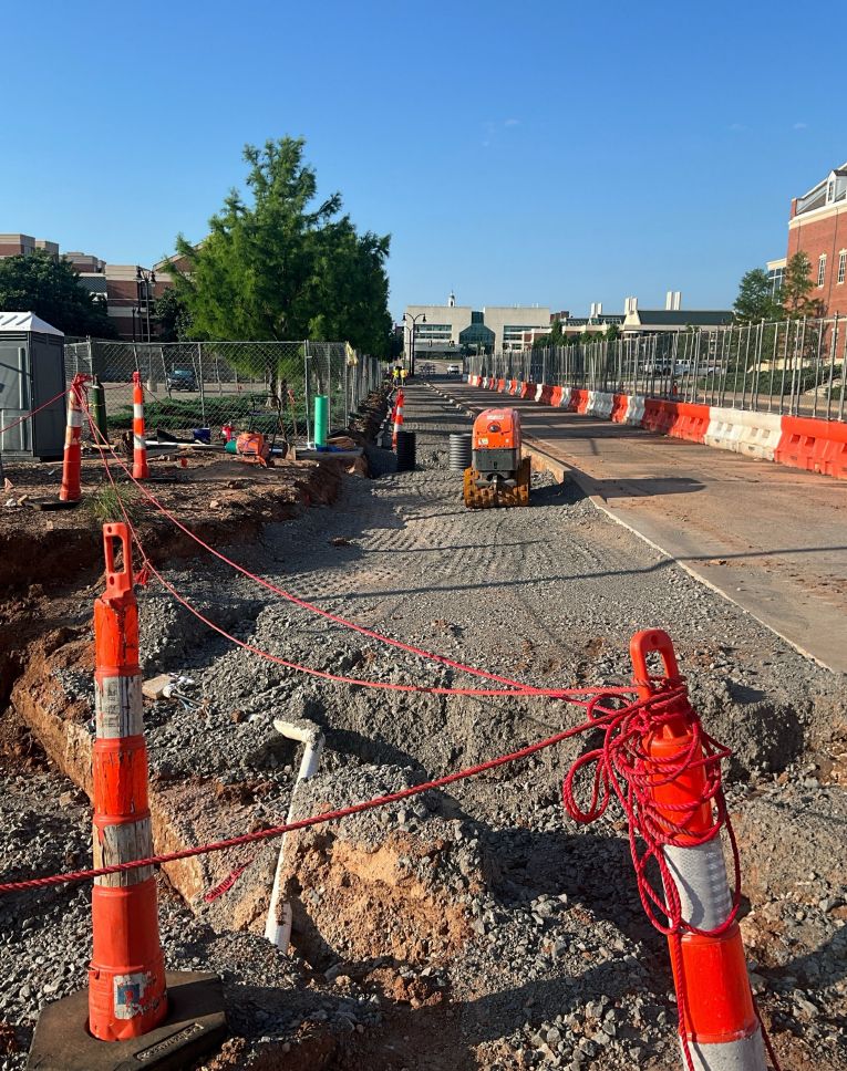 Fenced construction zone where a tamping machine compacts crushed rock for a sidewalk expansion.