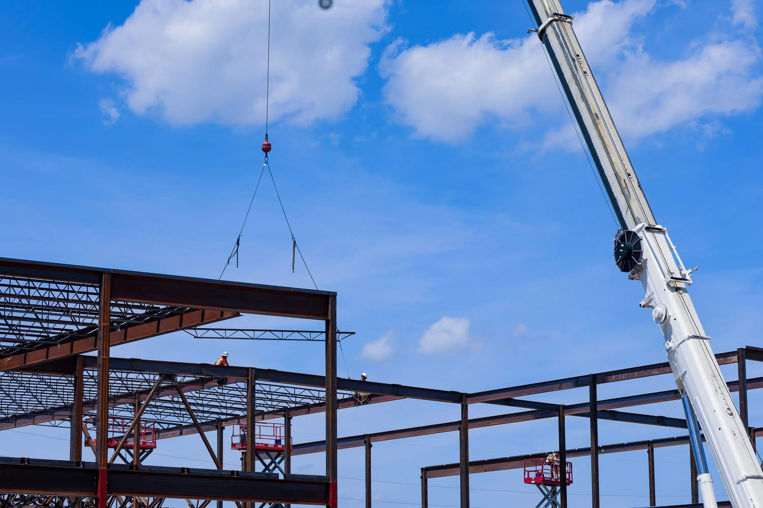 Construction workers install steel beams with a crane as the building frame rises against a blue sky.