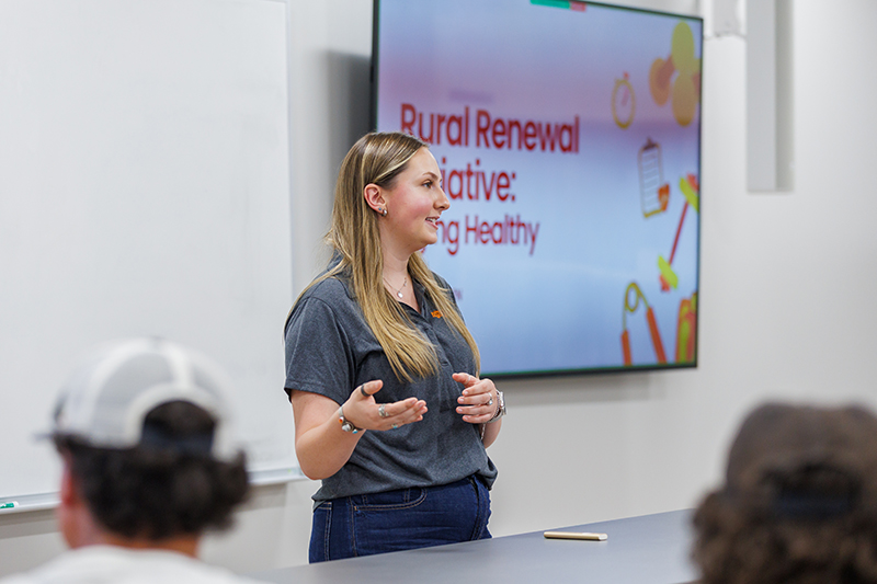 A presenter stands at the front of a classroom or meeting space, speaking to an audience seated in the foreground. Behind the presenter, a large display screen shows a slide with the title "Rural Renewal Initiative: Eating Healthy" and colorful nutrition-themed graphics. A whiteboard is mounted on the wall beside the screen, and a mobile phone rests on the table in front of the presenter.