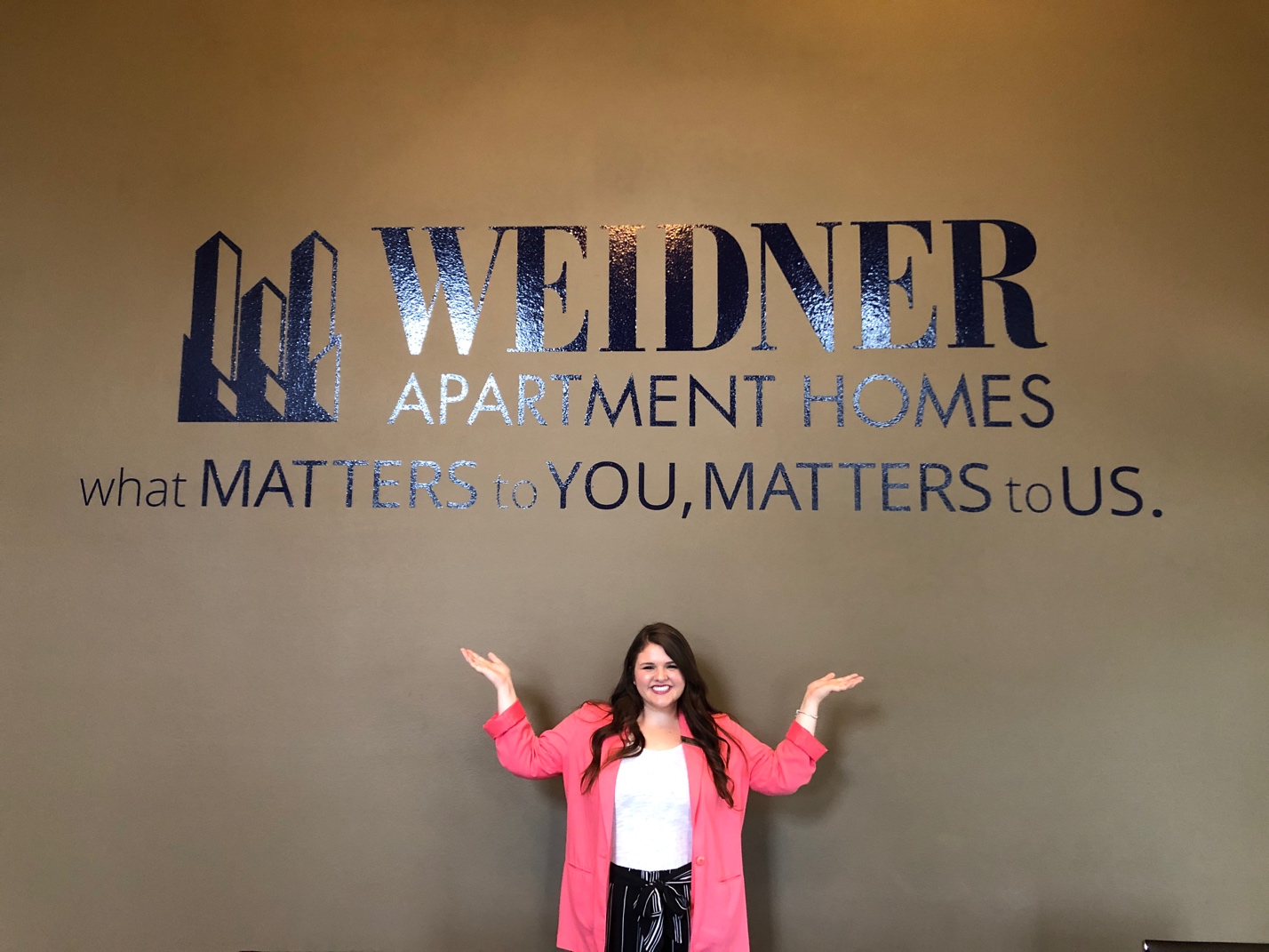 Female student wearing a pink blazer poses with a smile in front of a wall that says "Weidner Apartment Homes"