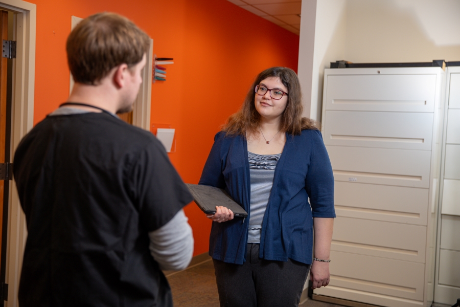 Female with papers in hand standing and talks to a man with his back to the camera. the man is wearing scrubs.